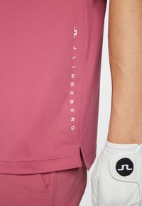 Kortärmad rosa polotröja med sidospringor, prydd med vit logotyp längs fållen och en strukturerad design. I sällskap med en vit golfhandske.