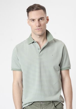 Hombre con camiseta polo de manga corta a rayas verdes claras y blancas con cuello levantado, de pie sobre fondo blanco liso.