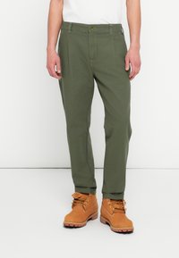 Oliwkowe spodnie chino z bocznymi kieszeniami, zapięciem na guziki i zwężanymi nogawkami, zestawione z jasnobrązowymi butami roboczymi na sznurowadła i z teksturowanymi podeszwami.