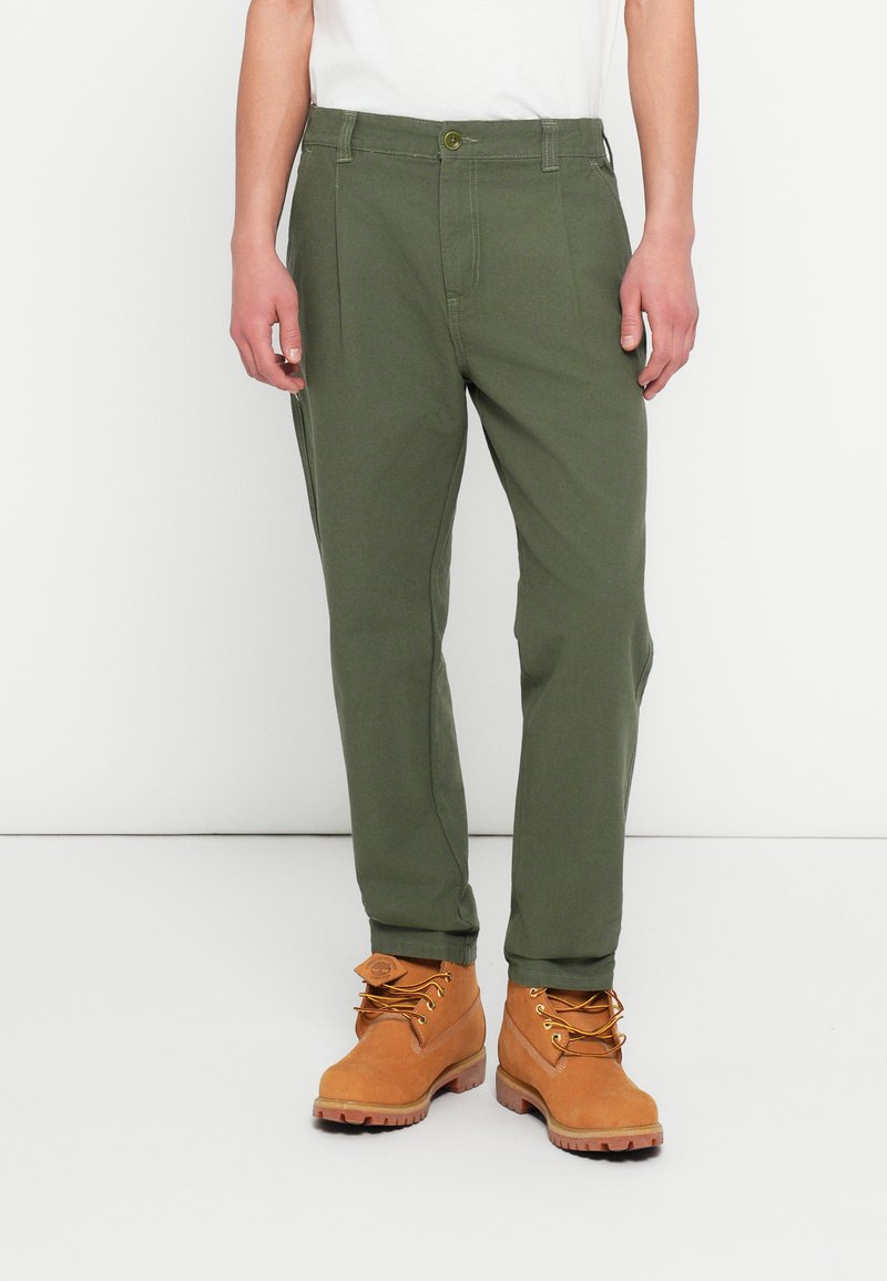 Oliwkowe spodnie chino z bocznymi kieszeniami, zapięciem na guziki i zwężanymi nogawkami, zestawione z jasnobrązowymi butami roboczymi na sznurowadła i z teksturowanymi podeszwami.