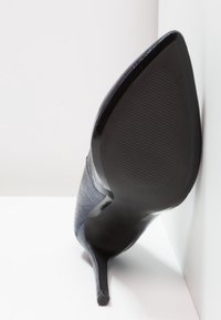 Chaussure à talon haut noire avec un dessus en cuir texturé, bout pointu et un talon fin et élégant. La semelle présente un motif en relief pour une meilleure adhérence.