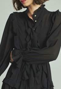 Vestido negro de chiffon con detalles con volantes, mangas largas con puños elásticos y botones decorativos en el escote. Tejido texturizado.