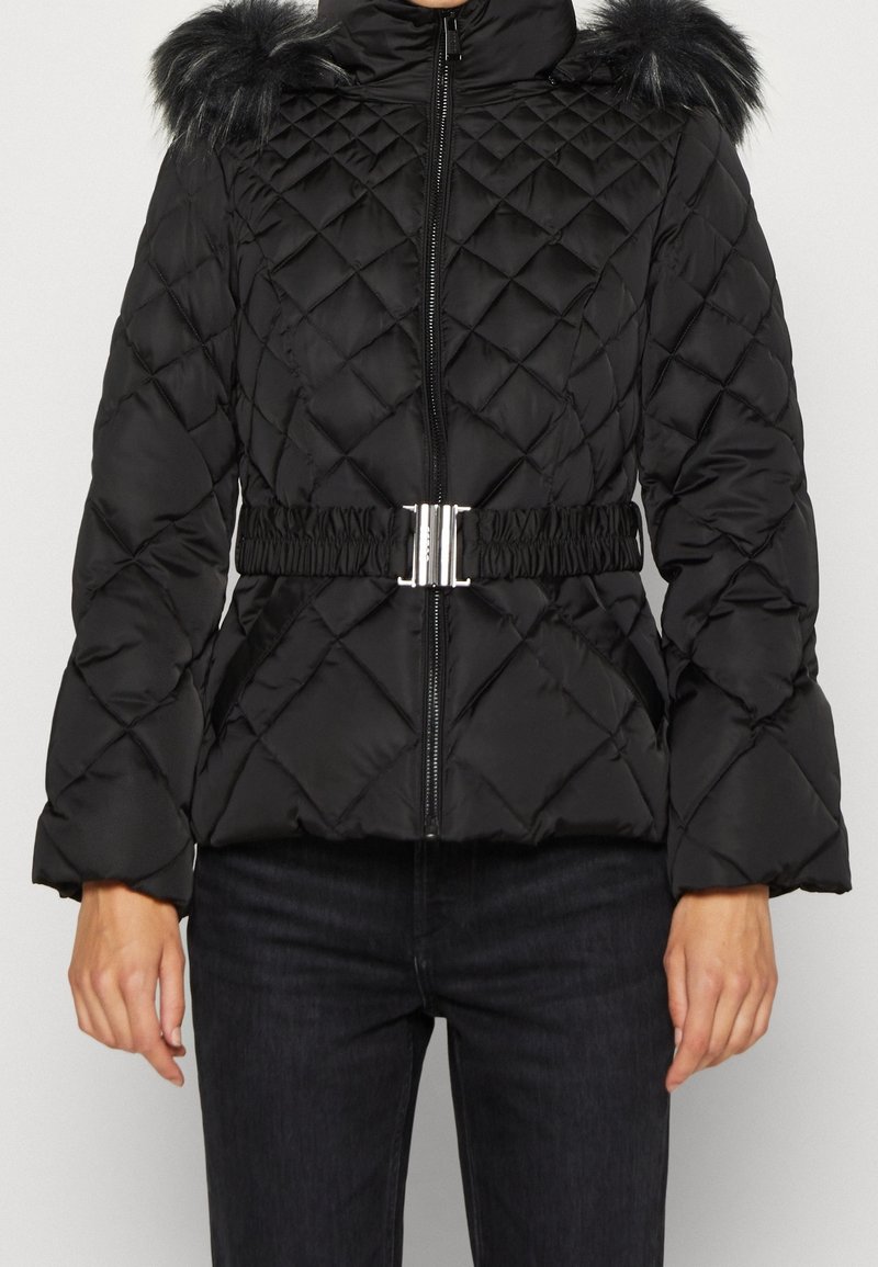 Veste matelassée noire avec capuche bordée de fourrure, ceinture à boucle argentée à la taille et fermeture éclair à l'avant, portée avec un pantalon foncé.