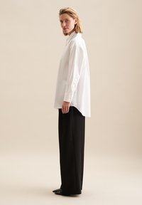 Witte overhemd met lange mouwen en hoge kraag, met knoopmanchetten, gecombineerd met zwarte wijde broek en puntige zwarte schoenen.