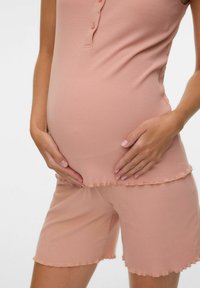 Femme enceinte portant un haut sans manches côtelé rose et un short, les mains caressant doucement son ventre sur un fond blanc.