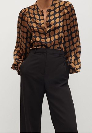 Femme portant un chemisier noir à gros pois dorés rentré dans un pantalon noir taille haute à jambes larges, mains dans les poches.