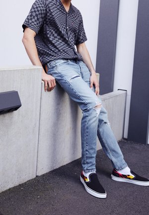 Person trägt ein kurzärmeliges kariertes Hemd, zerrissene hellblaue Jeans und schwarze Slip-On-Schuhe mit Flammenmuster und sitzt auf einer Betonmauer.