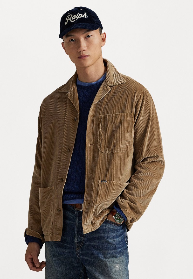 Mann som har på seg en marineblå caps, lysebrun cordfløyelsjakke med frontlommer, blå genser, jeans og et grønt klokke på håndleddet, står mot en hvit bakgrunn.