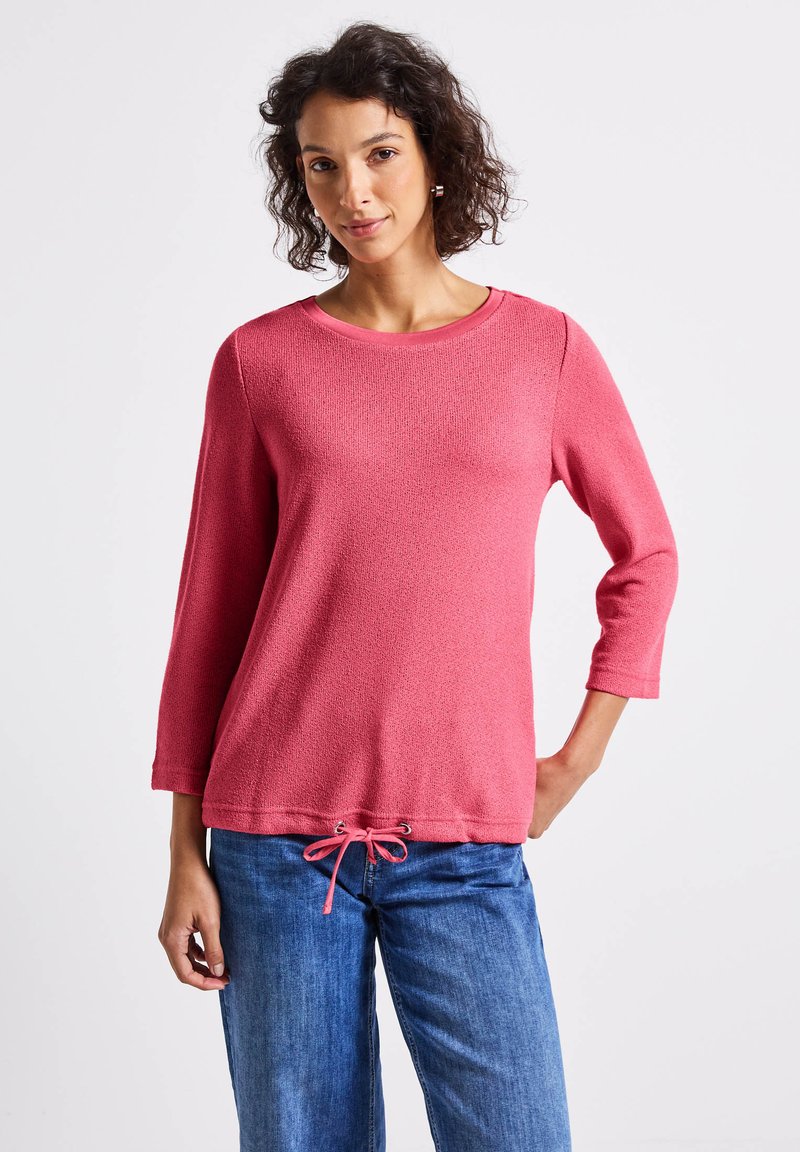 Rosa strukturiertes Sweatshirt mit Dreiviertelärmeln und Kordelzugdetails am Saum, kombiniert mit blauen Jeans.