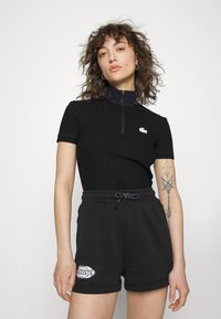 Top preto canelado de mangas curtas com gola de meio fecho, combinado com calções pretos com um patch do logótipo Lacoste e uma cintura ajustável.