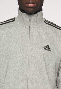 Siva zadrževena majica z zadrgo, izdelana iz mehkega materiala. Ima črn logo Adidas in tri črne črte na rami. Gladka tekstura, zasnova z visokim ovratnikom.