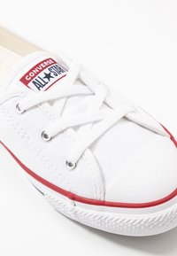 Bijela platnena tenisica s crvenim gumiranim potplatom i obrubom. Ima bijele vezice, okrugli vrh i zakrpu s natpisom "Converse All Star."