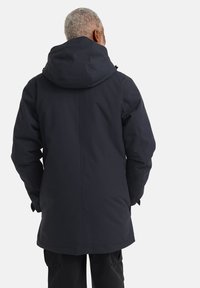 Parka bleu foncé avec capuche, texture douce et design ajusté. Présente des fermetures éclair et boutons, avec un ourlet arrière plus long pour une meilleure couverture.