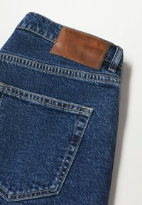 Jeans de mezclilla azul oscuro con un parche de cuero marrón en la cintura, que presentan un diseño tradicional de cinco bolsillos y costuras visibles.