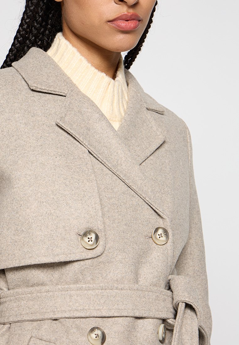Trench-coat en mélange de laine gris clair avec fermeture à double boutonnage, revers crantés et ceinture assortie. Pull crème à côtes en dessous.