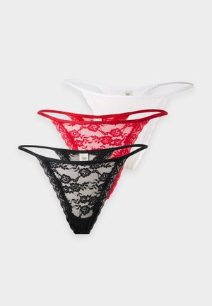 Femme aux longs cheveux foncés debout, portant un soutien-gorge en dentelle noire et une culotte assortie, sur un fond clair uni.