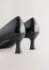 Paire d'escarpins noirs en cuir verni avec talons kitten et bouts pointus sur un fond clair et neutre.