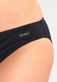 Parte inferior de bikini negra hecha de un material suave, con un acento de logo en tono dorado y un diseño simple y minimalista.