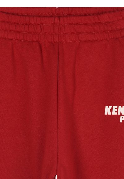 Un pantalon de survêtement rouge avec une taille élastique, fabriqué en tissu doux. Présente un texte blanc au niveau inférieur indiquant "KEN" et "P".