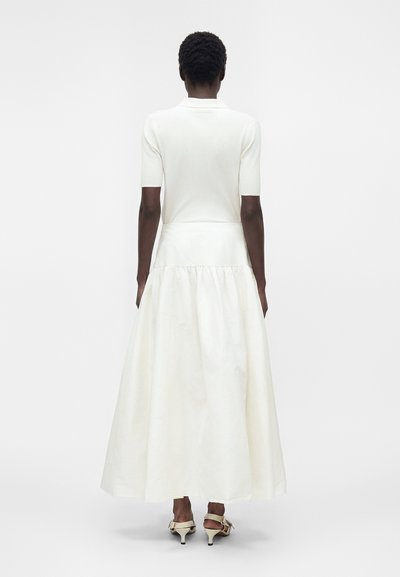 Personne portant un haut côtelé blanc à manches courtes et une longue jupe blanche fluide avec des sandales beiges à talons, debout de dos.