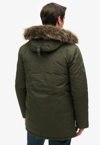 Parka verde oliva con capucha forrada de pelo sintético, tela texturizada, correa ajustable en la capucha y un parche redondo en la manga izquierda.