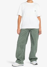 T-shirt en coton blanc avec une poche poitrine, associé à un pantalon large en velours côtelé vert olive, complété par des baskets claires.