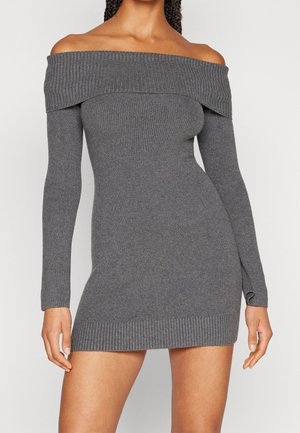 Kobieta w dopasowanej, długorękawowej, odsłaniającej ramiona, prążkowanej szarej mini sukience na jednolitym tle.