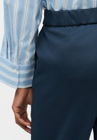 Κοντινό πλάνο στο χέρι ενός ατόμου δίπλα σε ναυτικό μπλε παντελόνι ψηλής μέσης και μανσέτα μακρυμάνικου ριγέ ανοιχτό μπλε πουκαμίσου με λευκό κουμπί.