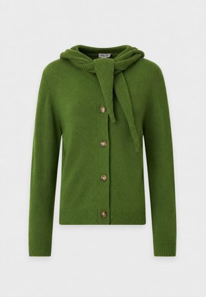 Femme portant un pull vert boutonné avec un détail d'écharpe, un jean large bleu clair et des chaussures noires, debout avec une main dans la poche.
