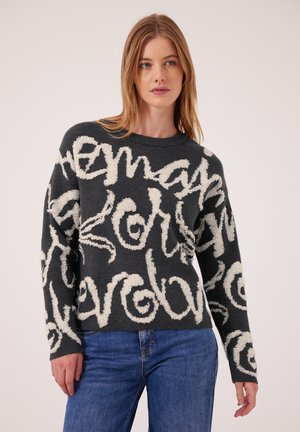 Kvinde iført en mørkegrå sweater med store hvide abstrakte scriptmønstre og blå jeans mod en ensfarvet baggrund.
