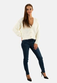 Cardigan bianco e peloso con scollo a V e tre bottoni, abbinato a jeans skinny blu scuro e tacchi neri. Design semplice e texturizzato.