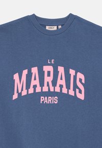 Modra sveternica z okroglim izrezom in rebrastimi manšetami, s poudarnim roza napisom "LE MARAIS PARIS" na sprednji strani.