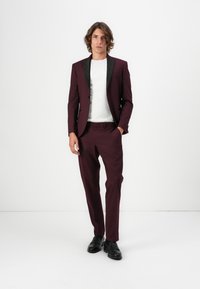 Bordeauxfarbener Anzug mit schwarzen Revers, taillierter Schnitt, Brusttasche und knöchellangen Hosen. Darüber ein weißes T-Shirt und schwarze Lederschuhe.