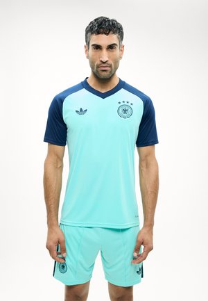 Férfi világoskék focimezben és rövidnadrágban, tengerészkék ujjakkal, egyszínű fehér háttér előtt áll, előre néz.