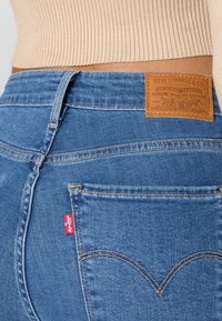 Gros plan sur la poche arrière bleue d'un jean Levi’s et sur l'écusson en cuir de la marque, portés avec un pull côtelé beige.