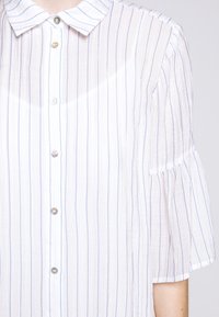 Blouse blanche semi-transparente à fines rayures bleues verticales, boutonnée sur le devant, manches courtes évasées et col pointu portée par une personne.