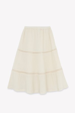 A cream-colored tiered skirt with an elastic waistband, featuring delicate mesh accents at the seams and a lightweight, textured fabric.