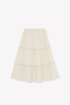 Krémová sukňa v tiered štýle s elastickým pásom, ozdobená jemnými sieťovanými detailmi na švoch a vyrobená z ľahkej textúrovanej látky.