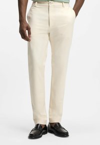 Mand iført cremefarvede bukser med lige ben med hænderne i lommerne og sorte læderloafers, vist fra talje til fødder.