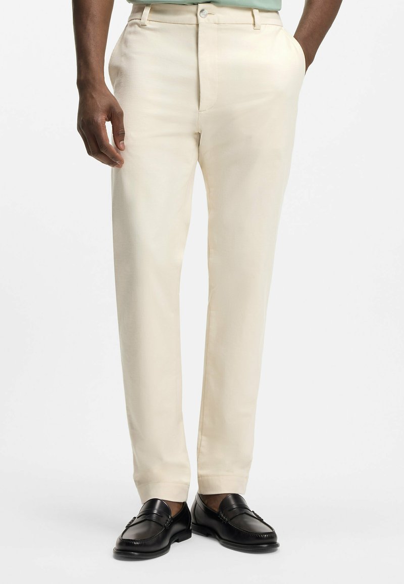 Mand iført cremefarvede bukser med lige ben med hænderne i lommerne og sorte læderloafers, vist fra talje til fødder.