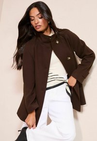 Rudas vilnos mišinio paltas su aukštu apykakliu, dvigubomis sagomis priekyje ir struktūruotomis rankovėmis. Dėvimas ant kūno spalvos palaidinės, derinamas su baltais kelnėmis.