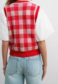 Czerwono-różowa, w kratkę, dziana kamizelka z czerwonym ściągaczem, w połączeniu z białą bluzką z bufiastymi rękawami i jasnoniebieskimi spodniami z wysokim stanem.