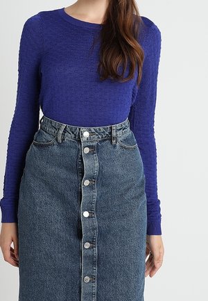 Mujer con un top morado texturizado de manga larga metido dentro de una falda vaquera azul de talle alto con botones en la parte delantera.
