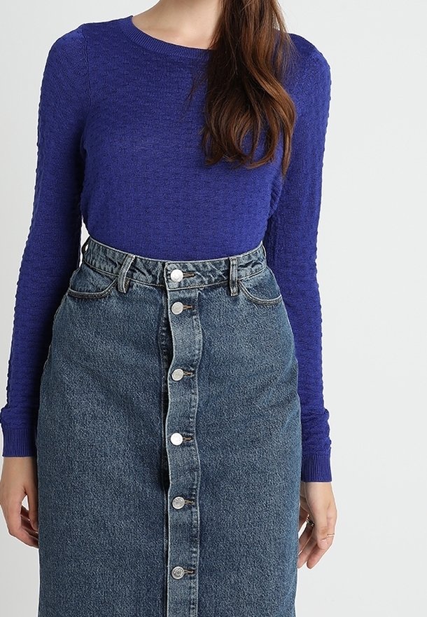 Femme portant un haut violet texturé à manches longues, rentré dans une jupe en jean bleue taille haute avec des boutons sur le devant.