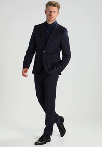 Tamsiai mėlynas kostiumas siauru kirpimu ir įpjovomis atlapuose, derinamas su tamsia palaidine. Baigtas blizgiais juodais batais su subtilia tekstūruota rašto detalė.