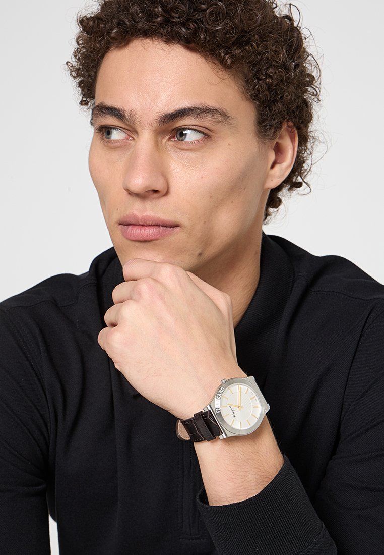 Jeune homme aux cheveux bouclés portant un haut noir, reposant son menton sur sa main, montrant une montre-bracelet en argent avec un bracelet en cuir marron.