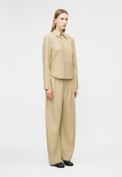 Žena stojaca v béžovej koženej dlhorukávovej košeli a širokých nohaviciach s čiernymi topánkami na bielom čistom pozadí.