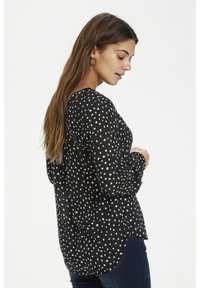 Schwarze Bluse mit weißen Polka-Dots, langen Ärmeln und abgerundetem Saum. Hergestellt aus leichtem Stoff mit lockerer Passform und subtiler glänzender Optik.