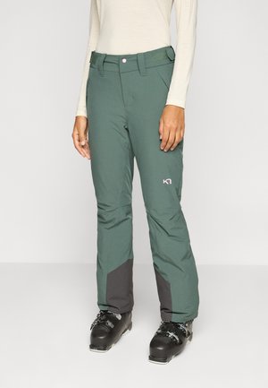 Femme portant un pantalon de ski vert avec des guêtres renforcées noires et des chaussures de ski noires sur un fond uni.
