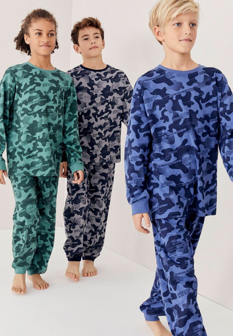 Tre børn går barfodede iført langærmede camouflage-nattøj i grønne, grå og blå toner mod en ensfarvet baggrund.
